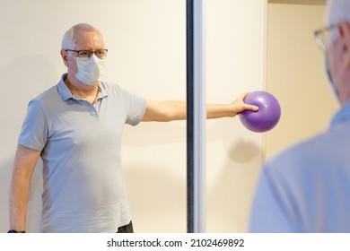 Older Man Doing Kinesiology Exercises To Recover From Shoulder Tendinitis, Looking In The Mirror With A Medicine Ball