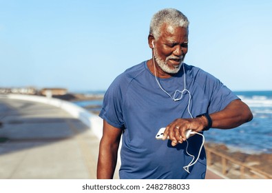 Older man enjoying a seaside workout, checking his fitness watch while listening to music for motivation. - Powered by Shutterstock