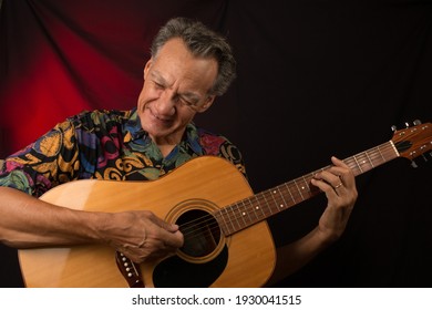 Older Man Having Fun Playing His Acoustic Guitar Against A Red Lit Background