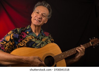 Older Man Having Fun Playing His Acoustic Guitar Against A Red Lit Background