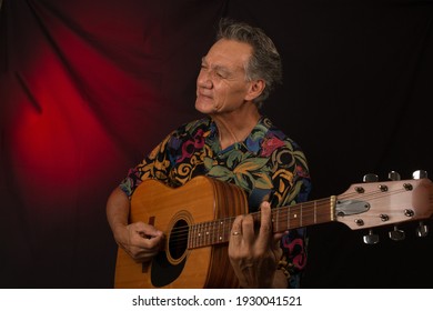 Older Man Having Fun Playing His Acoustic Guitar Against A Red Lit Background