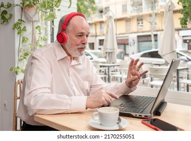 Older Man With Headphones Uses Laptop To Participate In Conference, Webinar, Sitting In Coffee Shop