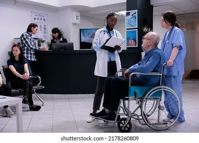 Older Man Living With Disability Being Helped By Caring Nurse Talking With Doctor Holding Clipboard With Patient History. Senior Citizen Using Wheelchair In Clinic Reception With Diverse People.