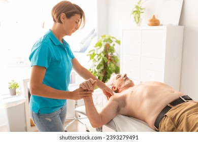 An older man, lying on the table in a physiotherapy clinic, during a manual therapy session with his physio, which assesses shoulder mobility - Powered by Shutterstock