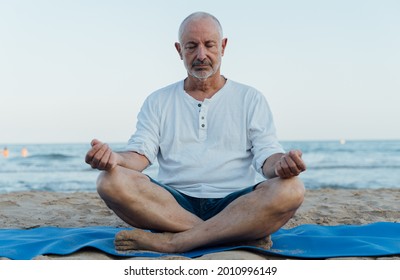 Older Man Meditating On The Beach On A Mat In The Sand