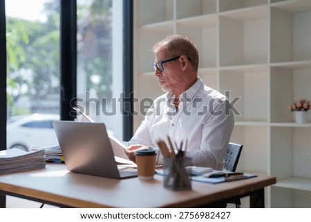Older man reviewing documents and using laptop at his modern workplace