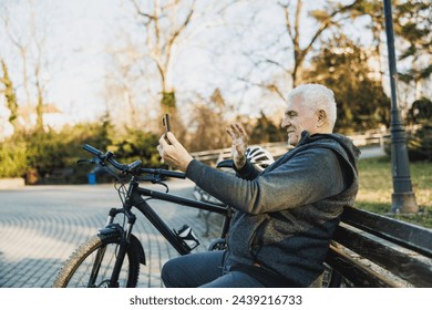 An older man is seated on a bench, engrossed in his cell phone. He making a video call in a sunny city park. - Powered by Shutterstock