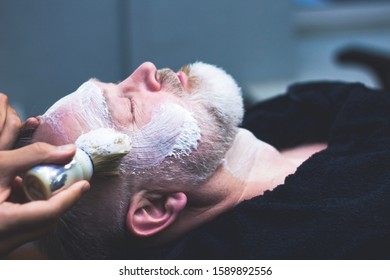 An Older Man Is Shaved In A Barber Shop. A Professional Worker Applies Shaving Foam To The Face Of A Client. Beauty And Care For Your Appearance