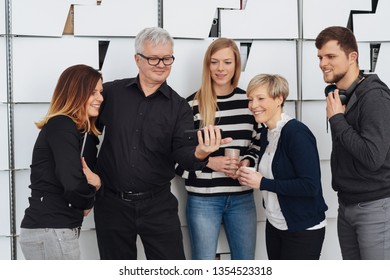 Older Man Showing Something To A Group Of His Friends As They Enjoy A Coffee Break At The Office Gathered Around Hime Looking At His Hand