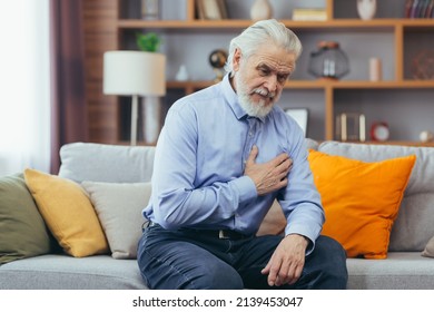 The older man sits at home alone, the grandfather holds his hands on his chest, his heart aches - Shutterstock ID 2139453047