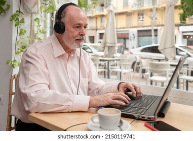 An Older Man Sitting In A Coffee Shop With Headphones Using His Laptop To Watch Online Classes And Webinar While Enjoying His Coffee