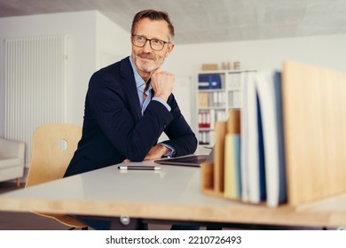 Older Man Sitting At Desk In Bright Room Working With Laptop Computer In Home Office. Mature Age, Middle Age, Mid Adult Casual Man In 50s, Confident, Thinking.