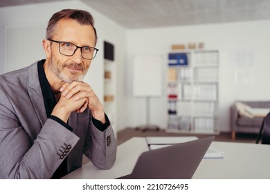 Older Man Sitting At Desk In Bright Room Working With Laptop Computer In Home Office. Mature Age, Middle Age, Mid Adult Casual Man In 50s, Confident, Thinking.