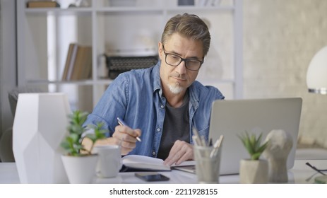 Older Man Sitting At Desk In Bright Room Working With Laptop Computer In Home Office. Mature Age, Middle Age, Mid Adult Casual Man In 50s, Serious, Focusing On Work.