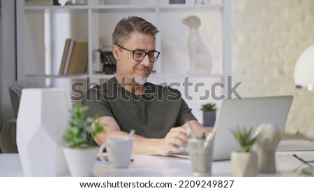 Older man sitting at desk in bright room working with laptop computer in home office. Mature age, middle age, mid adult casual man in 50s, confident happy smiling.
