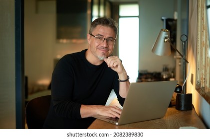 Older Man Sitting At Desk In Dark Cosy Room Working On Laptop Computer In Home Office. Mature Age, Middle Age, Mid Adult Casual Man In 50s, Confident Happy Smiling.