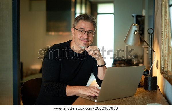 Older man sitting at desk in\
dark cosy room working on laptop computer in home office. Mature\
age, middle age, mid adult casual man in 50s, confident happy\
smiling.