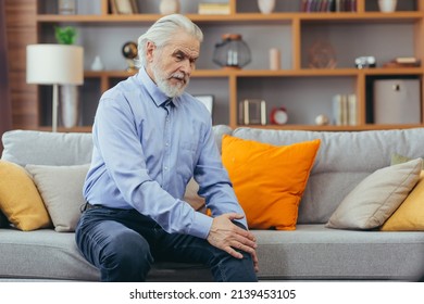The older man is sitting on the couch at home, has pain in the knee joint, holding his hands and feet