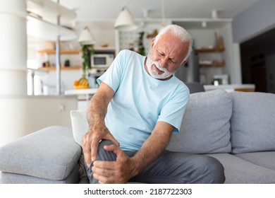 The Older Man Is Sitting On The Couch At Home, Has Pain In The Knee Joint, Holding His Leg, Osteoarthritis Concept.