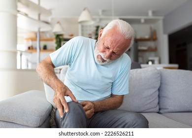 The Older Man Is Sitting On The Couch At Home, Has Pain In The Knee Joint, Holding His Leg, Osteoarthritis Concept.