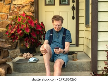 Older Man Sitting On Front Porch Of His House Talking On The Phone And Smiling On Nice Summer Evening In Midwest; Laptop Computer, Face Protecting Mask And Beverage Cup Next To Him 