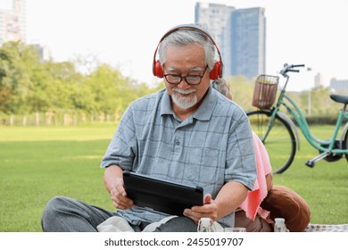 An older man is sitting on a grassy field, wearing headphones and holding a tablet. He is enjoying his time outdoors, possibly listening to music or watching a video. Concept of relaxation and leisure - Powered by Shutterstock