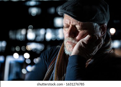 An older man is sitting outside at night. He is tired, rests his head on one hand and sleeps. City lights are in the background. - Powered by Shutterstock