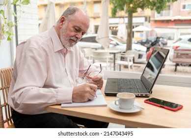 An Older Man Sitting At A Table With His Laptop In Front And Taking Notes Of His Business Finances In His Notebook, Coffee Cup And Mobile Phone Next To Him