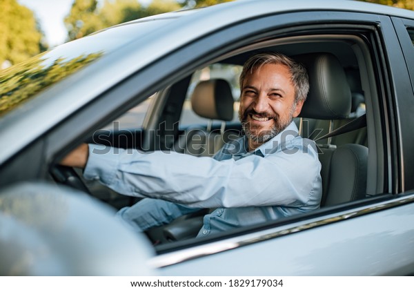 An older man smiling in the camera while he prepares\
to drive a car.