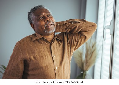 An Older Man Suffers From Back Pain. He Stands By The Window In The Living Room And Massages The Sore Spot With His Hand With A Sore Grimace On His Face. Elderly Man Suffering Pain Spine. 