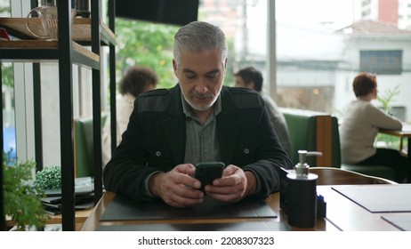 Older Man Using Cellphone Inside Coffee Shop Watching Content Online On Phone. A Middle Aged Person Looking At Smartphone Screen