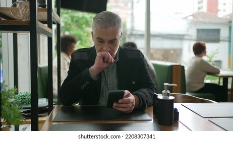 Older Man Using Cellphone Inside Coffee Shop Watching Content Online On Phone. A Middle Aged Person Looking At Smartphone Screen