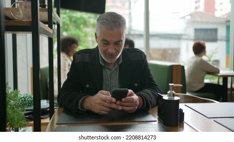 Older Man Using Cellphone Inside Coffee Shop Watching Content Online On Phone. A Middle Aged Person Looking At Smartphone Screen