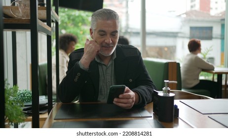 Older Man Using Cellphone Inside Coffee Shop Watching Content Online On Phone. A Middle Aged Person Looking At Smartphone Screen