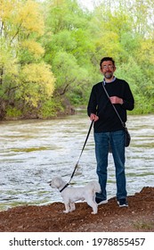 An Older Man Is Walking His Maltese Dog By The River