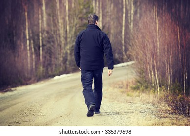 An Older Man Is Walking On The Road. He Is Walking Away. The Man Is Wearing Dark Jeans And A Black Jacket. Image Taken During Spring Time. Image Has A Vintage Effect Applied.