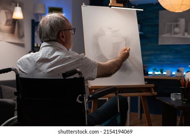 Older Man Who Is Paralyzed Shading Sketch In Light Study In Evening Class Feeling Inspired Drawing Vase Model On Paper. Focused Retired Art Teacher Using Wheelchair Doing Creative Artwork In Pencil.