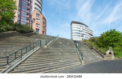 An Older Man And A Woman Climbing Together On A Wide And Tall Concrete Stairs On A Beautiful Spring Day. In The Background Are Colorful Tall Buildings.