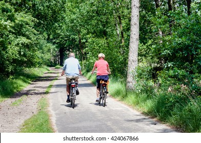 Older man and woman cycling together along a bike path through the forest. It's a warm day in summer.