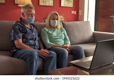 An Older Man And Woman Wearing Masks Sit Together On A Sofa With A Laptop In Front Of Them Talking To Family On A Video Call