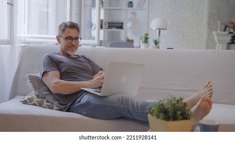 Older Man Working Online With Laptop Computer At Home Sitting On Couch In Living Room. Home Office, Browsing Internet. Portrait Of Happy, Mature Age, Middle Age, Mid Adult Man In 50s, Smiling.