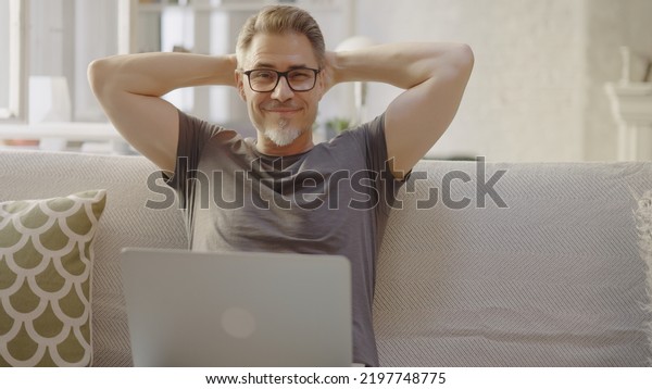 Older man working online with laptop computer at
home sitting on couch in living room. Home office, browsing
internet. Portrait of happy, mature age, middle age, mid adult man
in 50s, smiling.