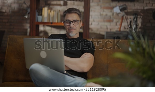 Older man working online with laptop computer at\
home sitting on couch in cosy living room. Home office, browsing\
internet. Portrait of happy, mature age, middle age, mid adult man\
in 50s, smiling.