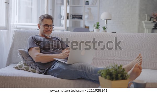 Older man working online with laptop computer at\
home sitting on couch in living room. Home office, browsing\
internet. Portrait of happy, mature age, middle age, mid adult man\
in 50s, smiling.