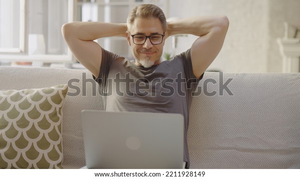 Older man working online with laptop computer at\
home sitting on couch in living room. Home office, browsing\
internet. Portrait of happy, mature age, middle age, mid adult man\
in 50s, smiling.