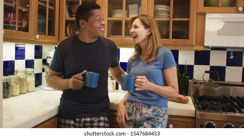 Older Married Couple Laughing While Chatting And Drinking Morning Coffee In Their Kitchen. Black And White Husband And Wife Talking About Their Day Wearing Pajamas