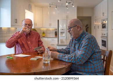 An Older Married Male Gay Couple Play A Card Game While Sitting At Their Kitchen Table. One Of The Men Is Taking A Drink Of Wine.