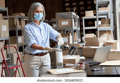 Older Mature Female Entrepreneur Small Business Owner Stock Worker Wearing Face Mask Using Sanitizer Cleaning Hands Preparing Working In Shipping Delivery Warehouse Office. Covid 19 Coronavirus Safety