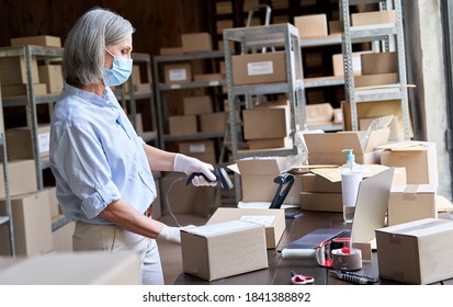 Older mature female online store small business owner worker wearing face mask packing package scanning postal drop shipping ecommerce retail order in box preparing delivery parcel in stock warehouse.