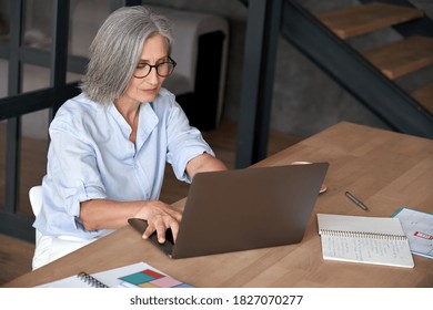Older Mature Middle Aged Business Woman Employee Using Laptop Typing Computer Sitting At Workplace Desk. Senior Old Lady 60s Grey-haired Businesswoman Entrepreneur Or Executive Working On Pc In Office
